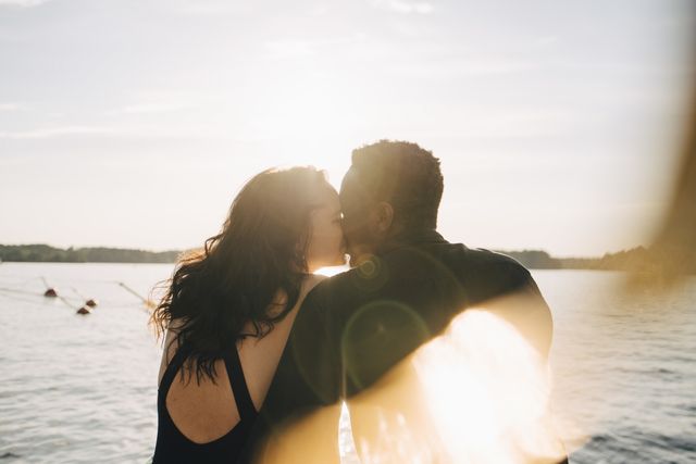 multi ethnic couple kissing while sitting against lake during summer