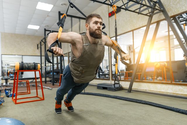 muscular bearded man dressed in military weighted armored vest doing exercises using straps systems in the gym sport, training, bodybuilding and healthy lifestyle concept