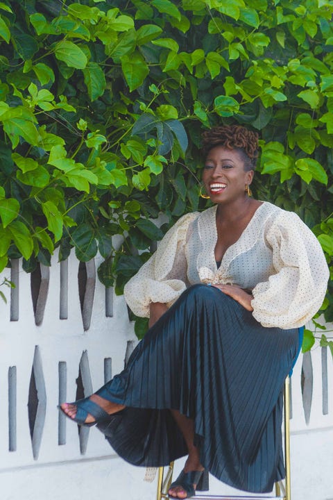 nana darkoa sekyiamah, author of the sex lives of african women, sits in a white blouse and navy pleated skirt in front of a white fence and a wall of leaves