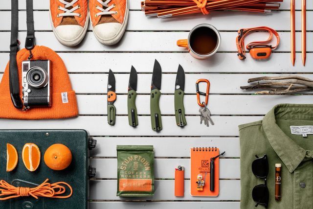 a flatlay photo of knives, a camera, camping gear, a shirt, sunglasses, and a cup of coffee