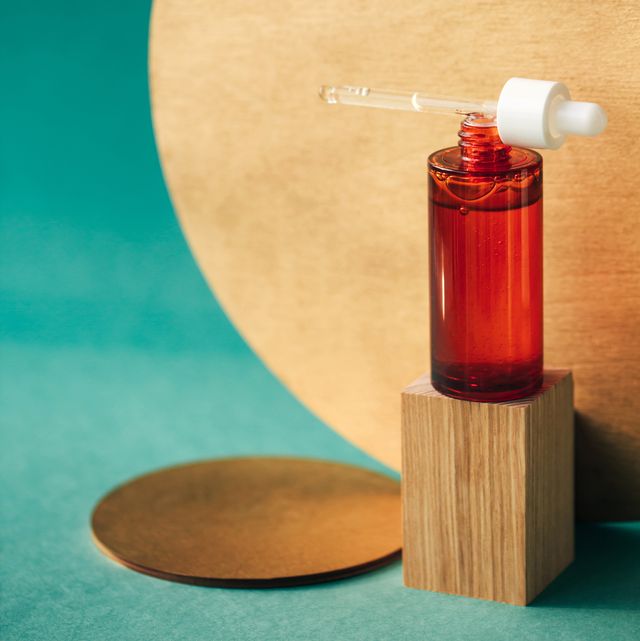 red glass bottle with pipette on it and with essential oil or moisturizing lotion on wooden podium near golden timber circles on table and on teal blue background concept of organic cosmetics and natural components front view with copy space for your design