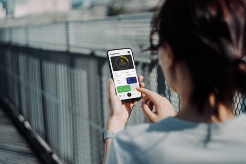 over the shoulder view of young asian sports woman using fitness app on smartphone to monitor her training progress while doing outdoor walk exercising in the fresh bright morning with shades of sunlight