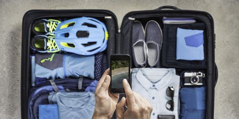 Overhead view of mans hands using smartphone touchscreen above packed suitcase with blue bike helmet, backpack, retro camera and shirt