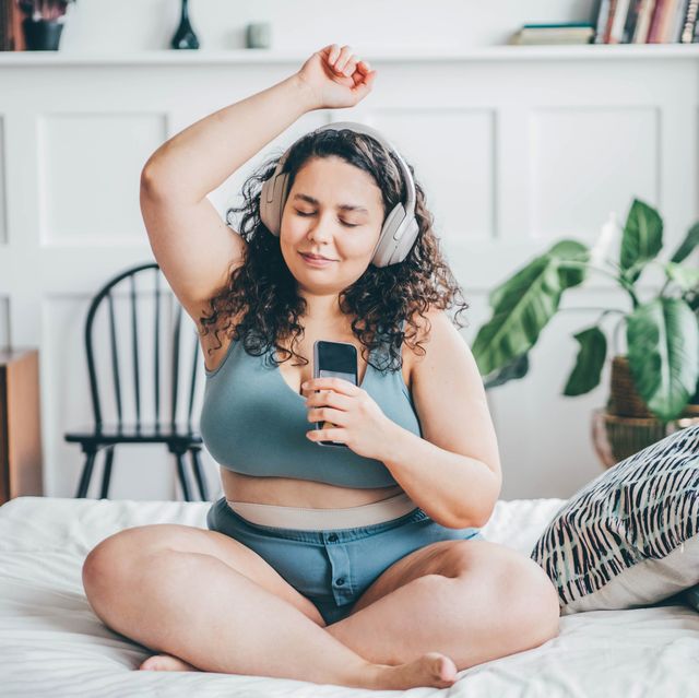 overweight young woman with loose hair in top and shorts listens to music with wireless headphones holding smartphone in hand and dances sitting on double bed