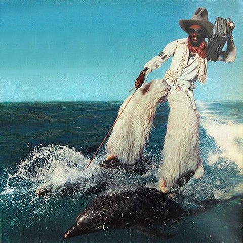 man carrying a boombox wearing furry chaps water skiing on two dolphins