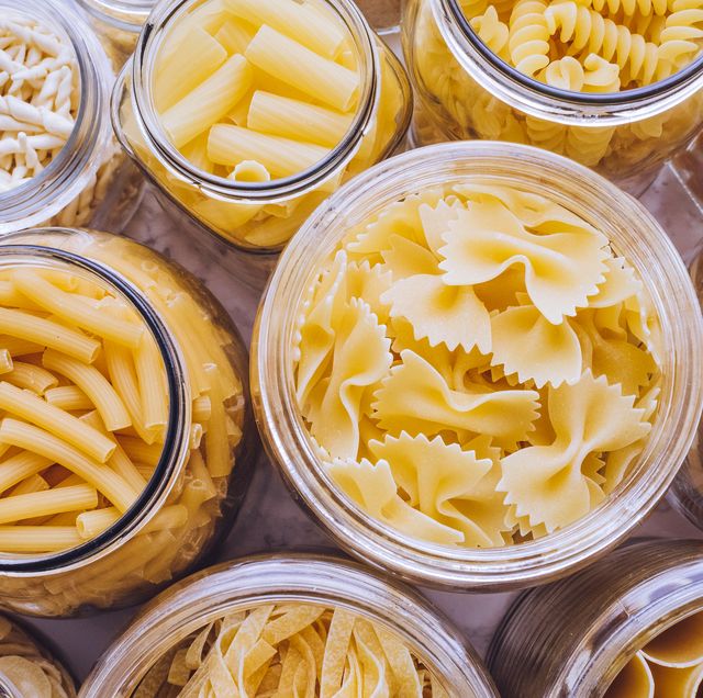 various types of pasta, including tube shapes, large shells, and bow tie shapes, in glass jars on marble background