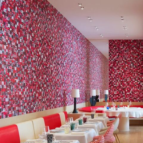 restaurant interior showing small tables in a line with white tablecloths ending in a large round pedestal table at the end with a banquette of alternative white and bright red leather or vinyl and a blue red and white amorphous checked wall