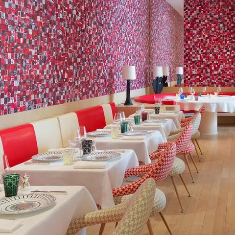 restaurant interior showing small tables in a line with white tablecloths ending in a large round pedestal table at the end with a banquette of alternative white and bright red leather or vinyl and a blue red and white amorphous checked wall