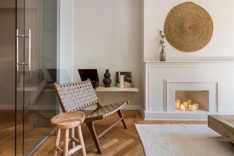 salón con chimenea blanca decorativa, decoración de pared de fibras naturales, butaca de piel trenzada y taburete de madera