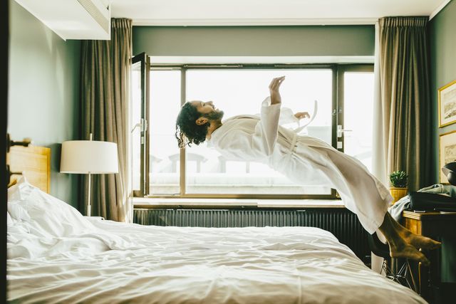 playful man wearing robe jumping on bed at hotel