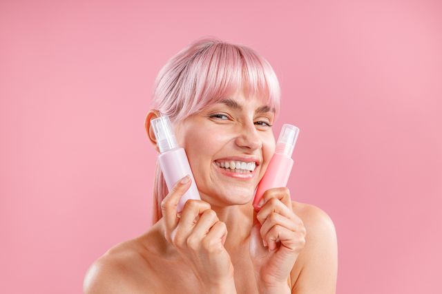 pleased young woman with pink hair holding two plastic spay bottles with beauty products near her face, posing isolated over pink background