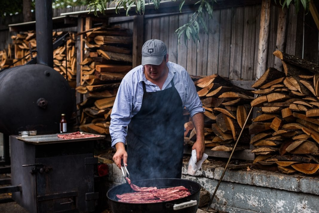 man grilling bacon with stacks of wood behind him