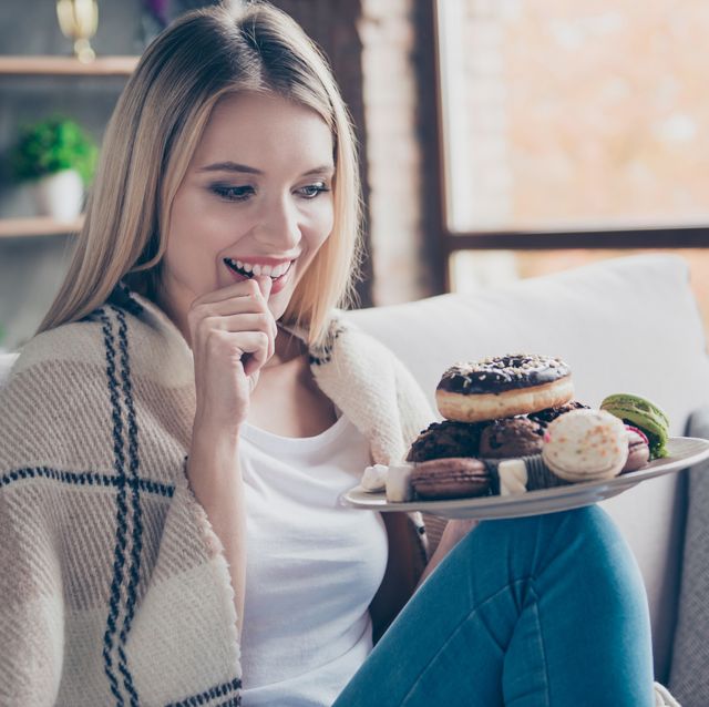 Portrait of beautiful emotional charming attractive sweet toothy woman sitting on sofa in living room, holding plate of donuts and macaroons, looking exciting satisfied