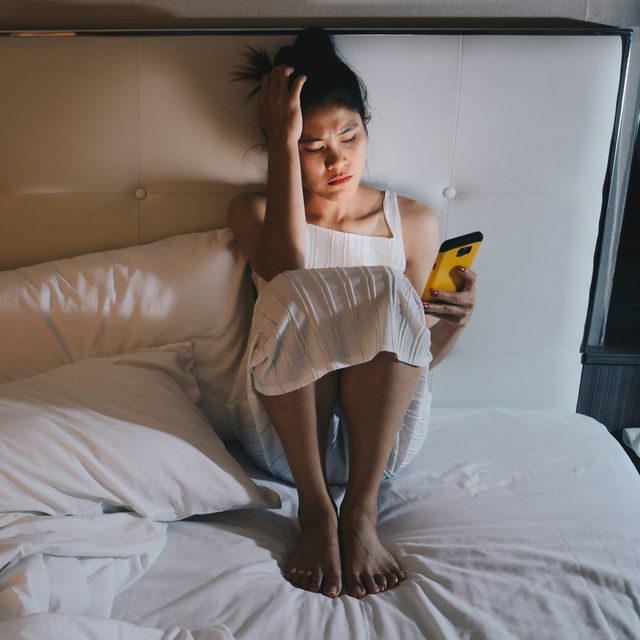 portrait of depressed woman sitting alone on the bed in the bedroom and looking to mobile phone in her hand