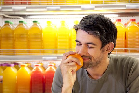 Portrait of smiling man sitting in front of fridge with rows of juice bottles in a supermarket smelling orange