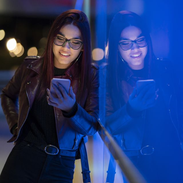 portrait of smiling teenage girl wearing leather jacket and glasses using smartphone at night