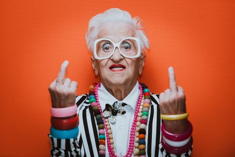 portrait of stylish senior woman wearing colorful jewelry showing middle finger against red background