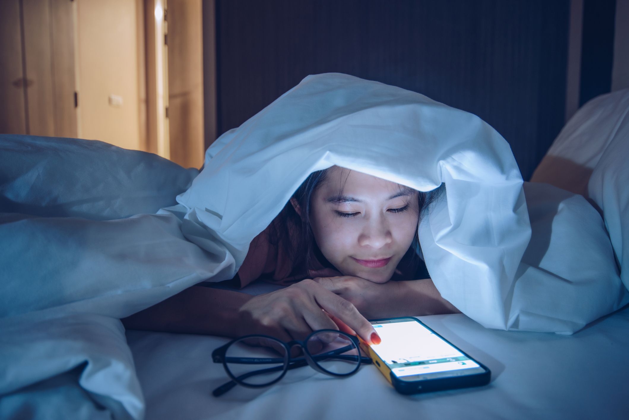 moters portretas, pasislėpęs po antklode, kalbantis ir naršantis internete išmaniuoju telefonu vėlai vakare lovoje