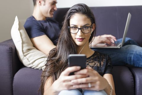 Portrait of young woman with cell phone and earphones and boyfriend on the couch in the background