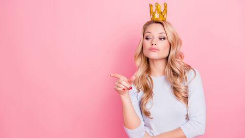 portrait with copyspace empty place of confident proud arrogant woman with gold crown on her head pointing forefinger, miss i want, isolated on pink background