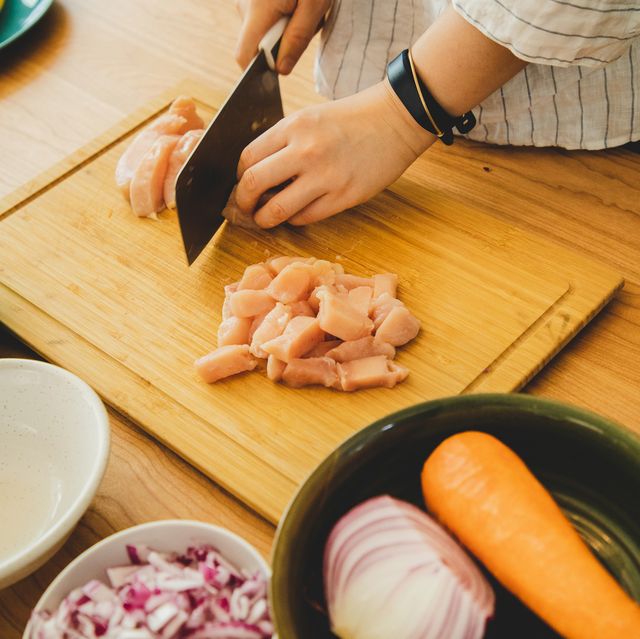 hands chopping raw chicken on a wooden cutting board with bowls of chopped vegetables nearby