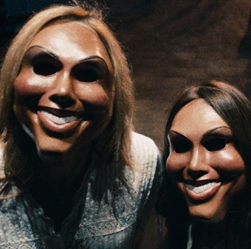 two menacing figures stand outside a door while wearing grinning masks in a scene from the purge, the first film if you want to watch the purge movies in order