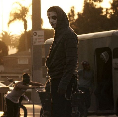 a man wears white face paint with a black, upside down cross painted on his forehead and stitches painted on his lips  in a scene from the purge anarchy, the second film if you want to watch the purge movies in order