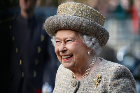 the queen smiles in a tweed hat and suit, wearing pearl earrings and necklace