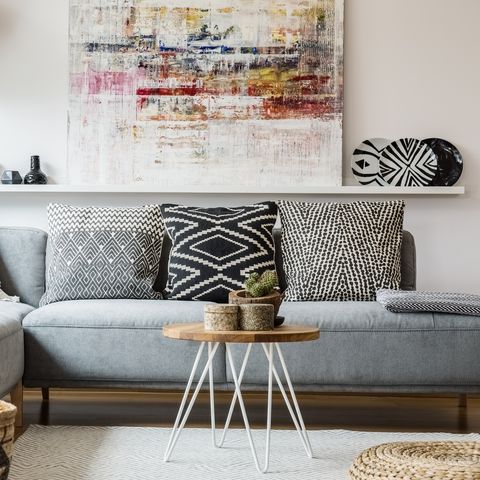 Cosy couch with cushions standing behind a small table and in front of a shelf with a painting in boho living room interior with baskets and white rug