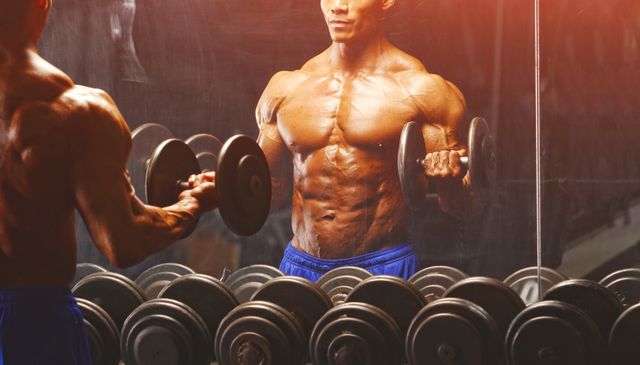 rear view of shirtless macho young man lifting dumbbells while looking in mirror
