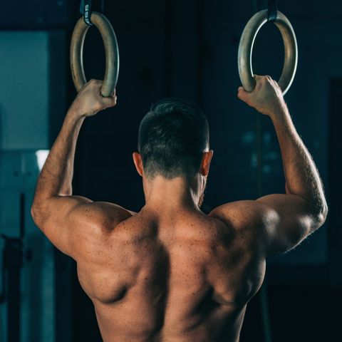 Rear View Of Shirtless Male Athlete Exercising On Gymnastics Rings In Gym