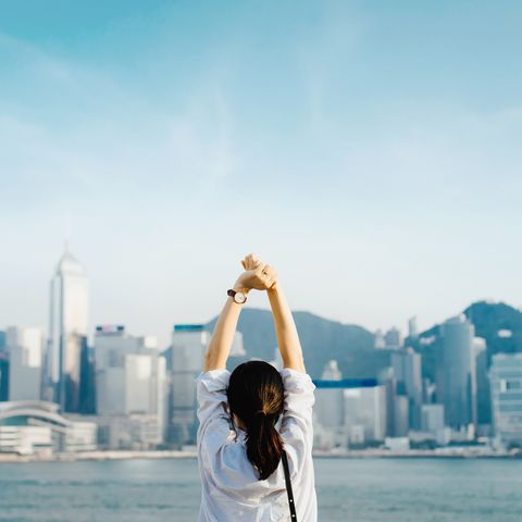 rear view of woman traveller enjoying her time in hong kong, taking a deep breath with hands raised against victoria harbour and city skyline