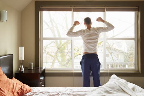 Rückansicht eines jungen Mannes, der sich vor dem Schlafzimmerfenster streckt