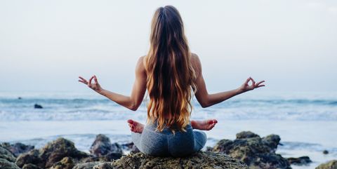 Rear view of young woman with long hair practicing lotus yoga pose on rocks at beach, Los Angeles, California, USA