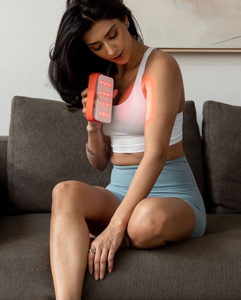 a woman sitting on a couch using a hand held red light therapy device on her arm