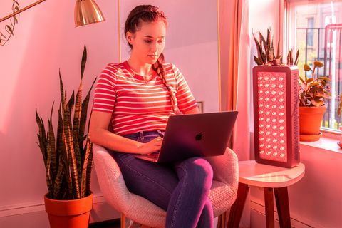 a woman working on her laptop with a red light therapy light on a table next to her