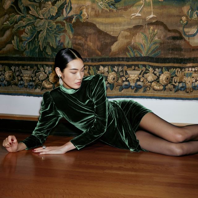 a model wears a velvet reformation dress in green in front of a tapestry to illustrate a roundup of the best velvet dresses to shop in 2021