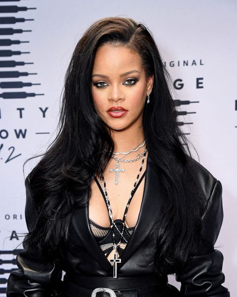 rihanna stands on the red carpet, wearing a leather jacket open over a black strappy fenty x savage bra and multiple cross shaped necklaces