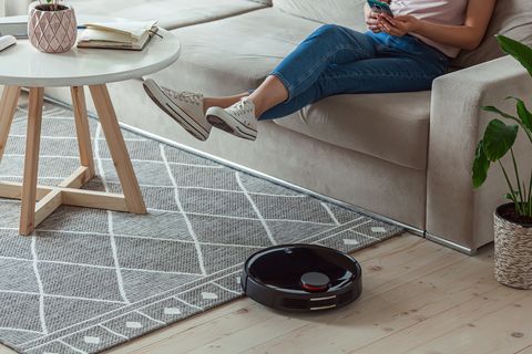 robot vacuum cleaner cleaning carpet, woman remote control mobile phone and enjoy rest, sitting on sofa home
