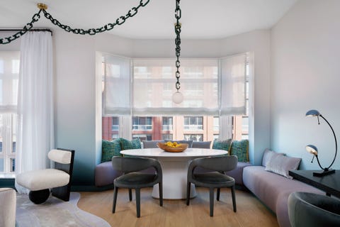 seating area with long curvaceous banquette in light purple fabric and lots of pillows set into the windowseat and then extending along the right wall and in front of the large curved window is a large white lacquer over pedestal table without two dark colored cut out chairs pulled up to it and a yellow bowl on the table and a pendant lamp on a long black chain coming in from the room on the left