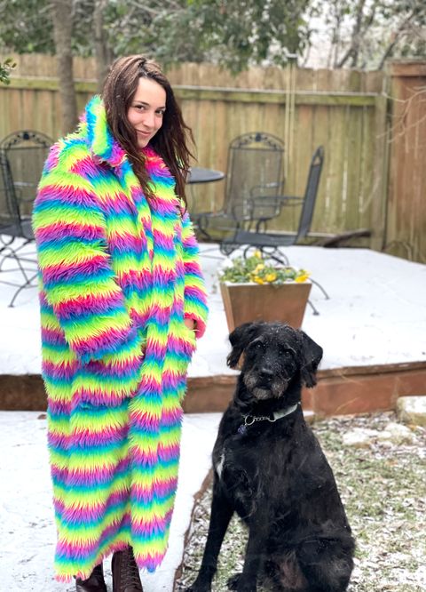 the author of the story rose minutaglio wearing her oversized technicolor coat with stripes of pink, green, yellow, and blue outside in austin, texas, when start started falling her 100 pound black hound dog stands next to her looking very cute
