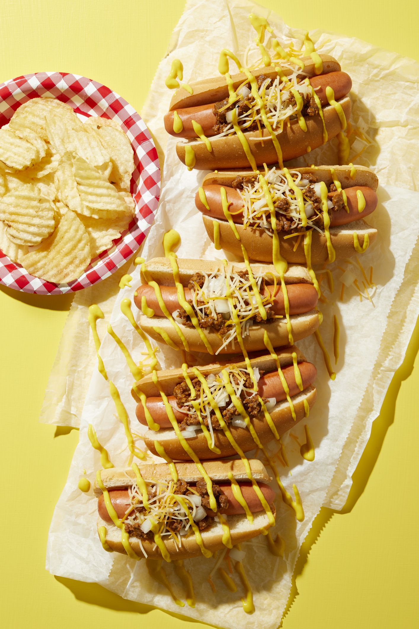 konusinių karštųjų šunų eilė su laistytomis garstyčiomis ir bulvių traškučių lėkštė geltoname fone, vaizdas iš viršaus