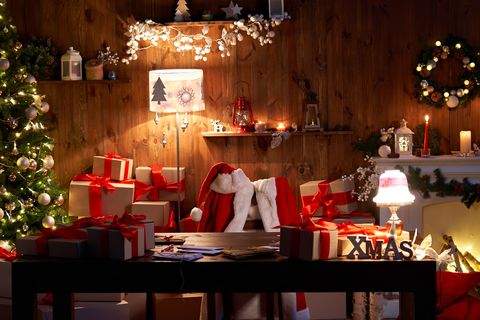 santa claus costume and hat hanging on chair at table with merry christmas decor gifts presents on holiday eve in cozy santa home workshop interior late in night with light on xmas tree and fireplace