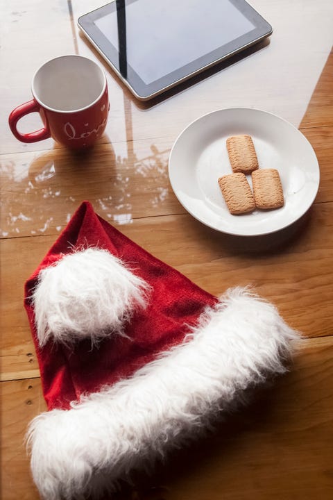 a santa hat on a wood table with a plate of cookies, a mug, and a tablet