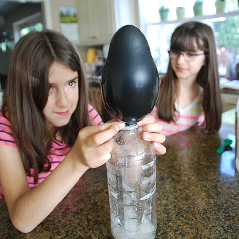 two girls watch a balloon inflating over a bottle as part of this at home science experiment for kids