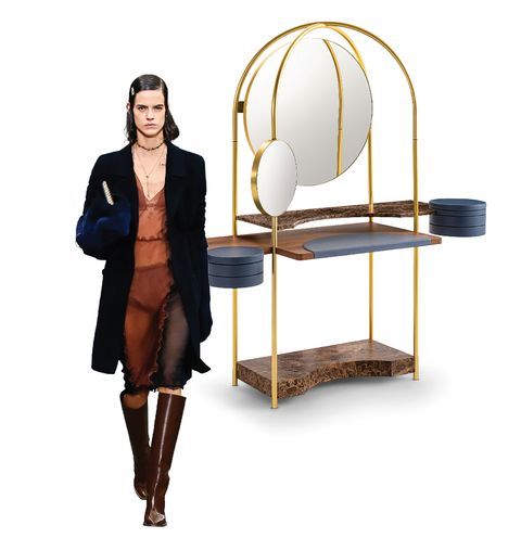 futuristic looking vanity with two round mirrors in a small and a large suspended in an open loop with spare wooden shelves beneath and next to it a model wearing a dark furry coat and orangey black outfit and tall brown leather boots
