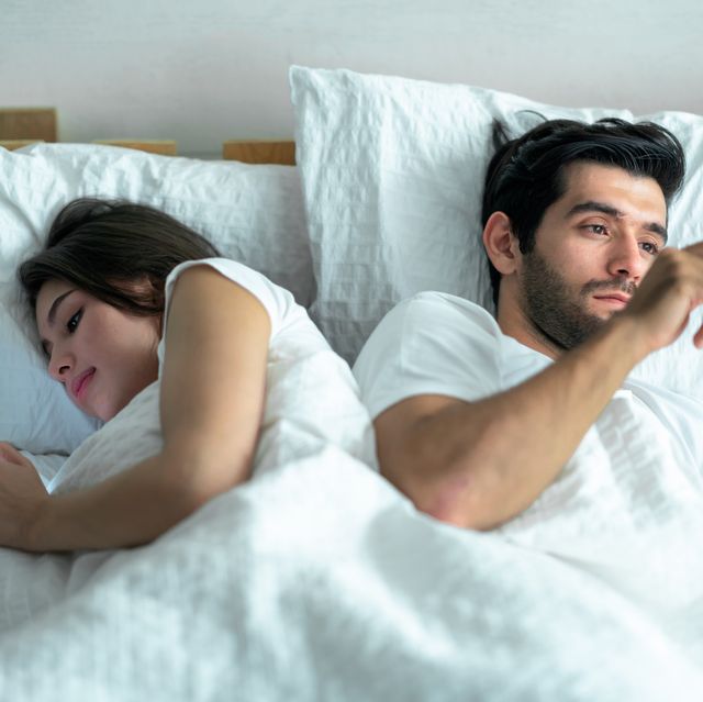 Shot of a young couple using their cellphones in bed at evening back to back .