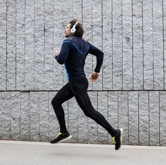 side view of male jogger wearing blue workout clothes running along concrete wall and listening to music through headphones