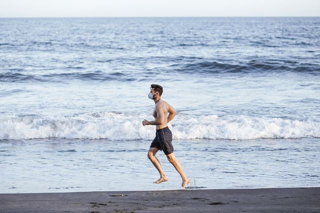 side view of shirtless mid adult man wearing face mask while running at beach during sunset