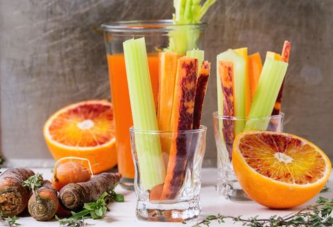 Sliced colorful raw carrots and celery as vegetarian snack, blood oranges and thyme with glass cup of fresh orange and carrot juice over white stone table.